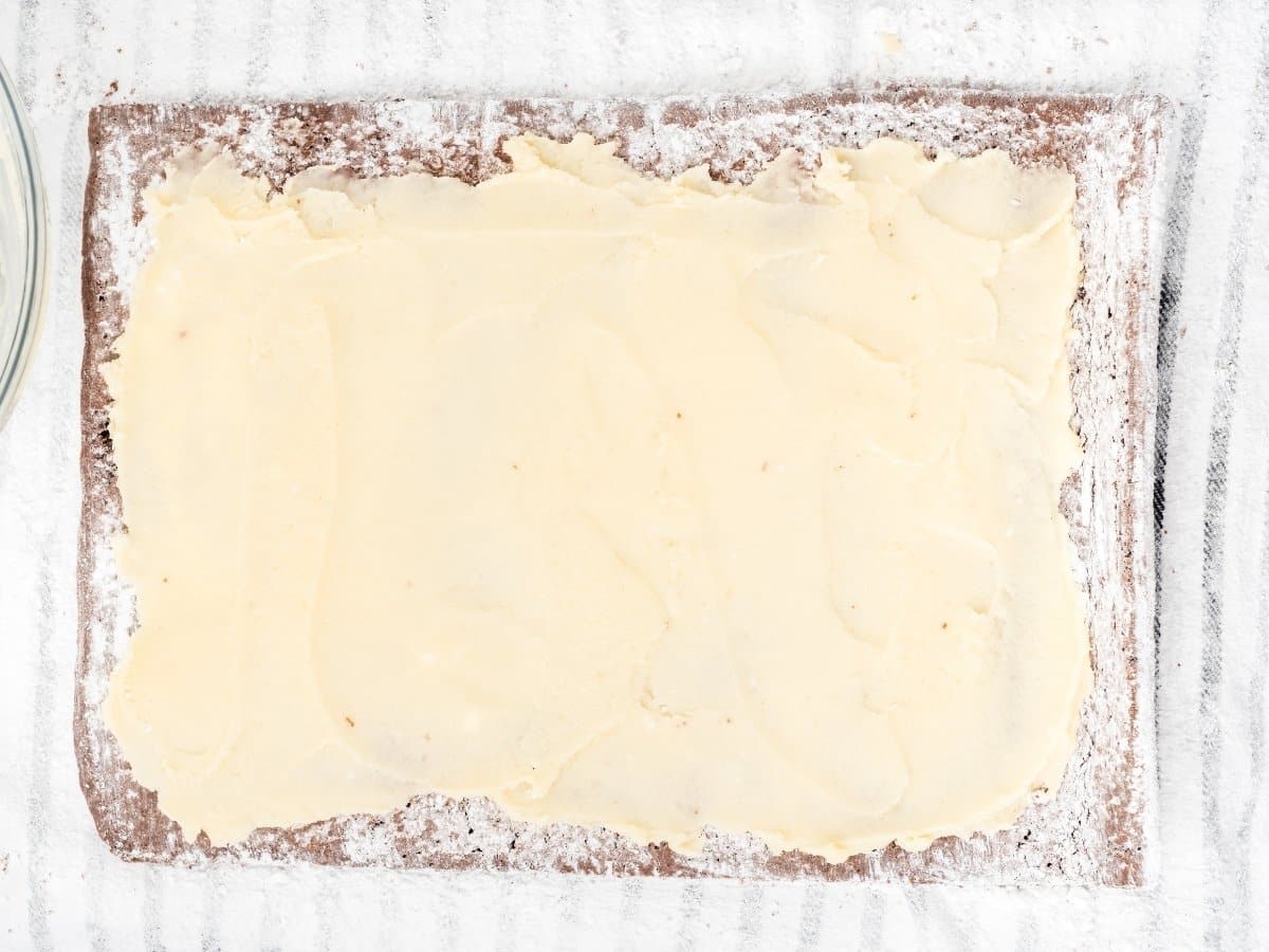 Top view of a chocolate sponge cake unrolled on a powdered sugar&ndash;dusted surface, with a layer of ermine filling spread evenly across the center, leaving a small border around the edges.