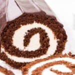 Chocolate Swiss roll cake sliced to show a tight spiral of fluffy chocolate sponge wrapped around a thick layer of smooth white cream filling. The cake is coated in a glossy chocolate ganache, with clean edges and a moist, tender crumb visible in each slice, set on a light surface with a soft, neutral background.