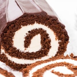Chocolate Swiss roll cake sliced to show a tight spiral of fluffy chocolate sponge wrapped around a thick layer of smooth white cream filling. The cake is coated in a glossy chocolate ganache, with clean edges and a moist, tender crumb visible in each slice, set on a light surface with a soft, neutral background.