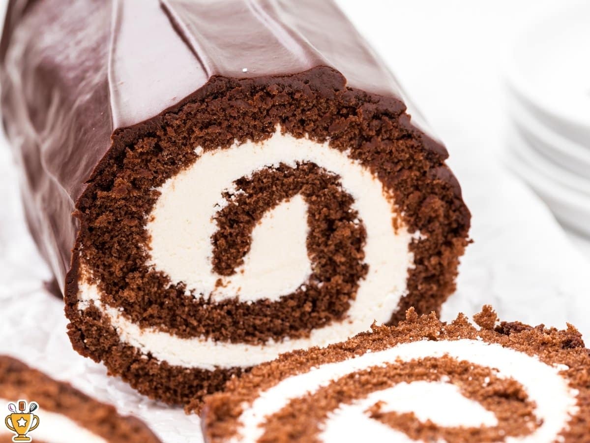 Chocolate Swiss roll cake sliced to show a tight spiral of fluffy chocolate sponge wrapped around a thick layer of smooth white cream filling. The cake is coated in a glossy chocolate ganache, with clean edges and a moist, tender crumb visible in each slice, set on a light surface with a soft, neutral background.