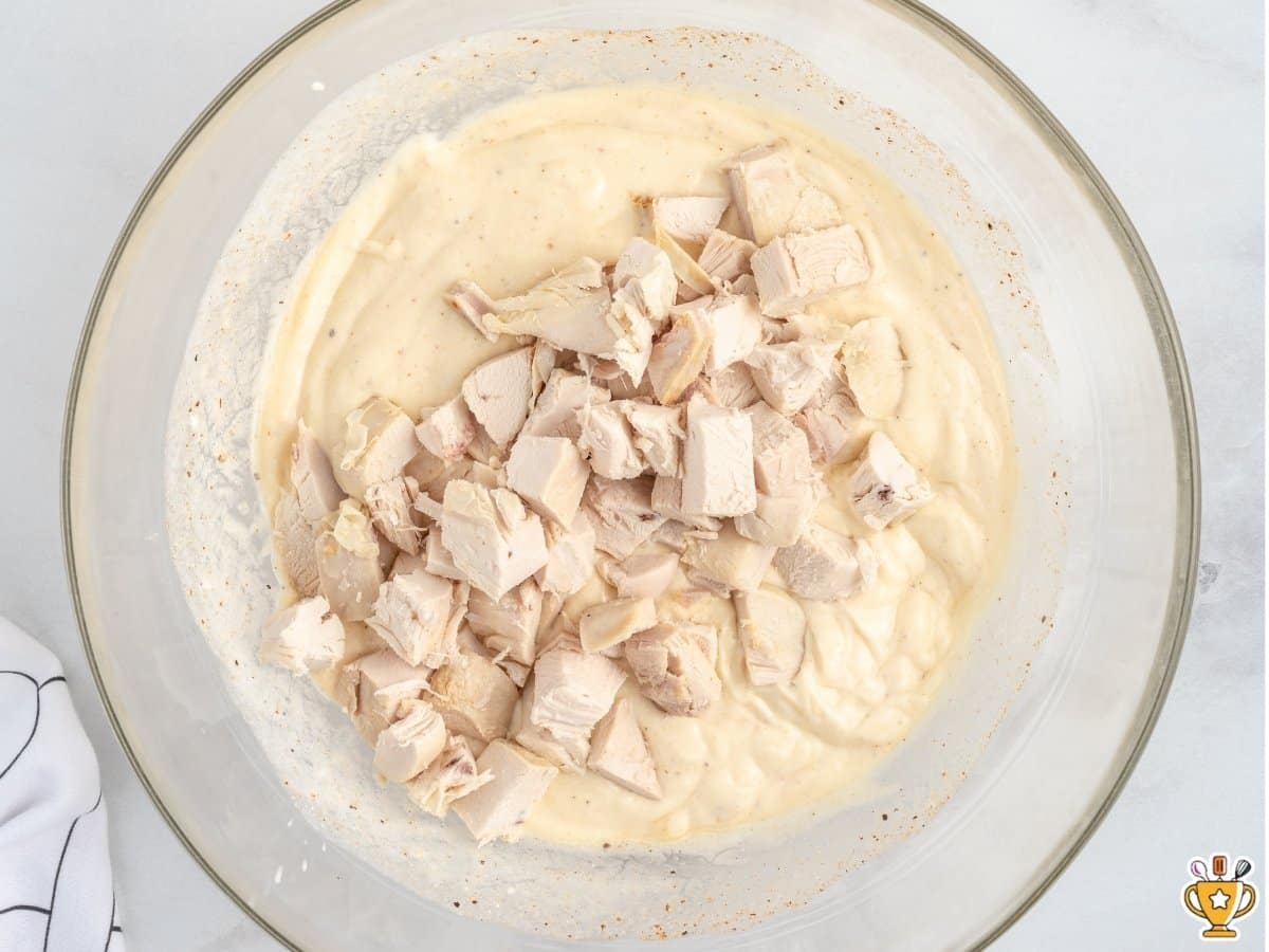 Diced rotisserie chicken being added to a bowl of creamy chicken casserole filling, with crushed Ritz crackers and additional chicken visible in bowls nearby.