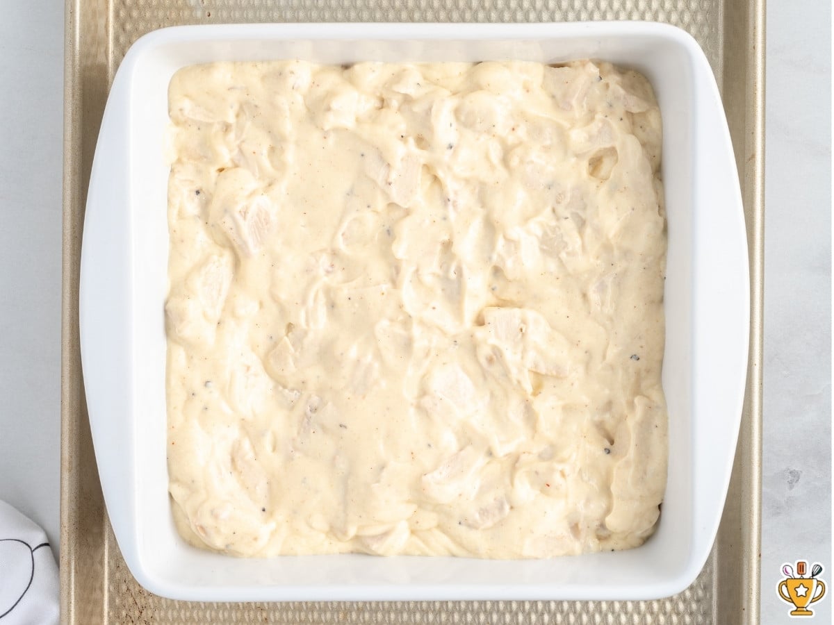 Creamy chicken casserole filling spread evenly in a white 8x8 baking dish, ready to be topped with buttery Ritz cracker crumbs before baking.