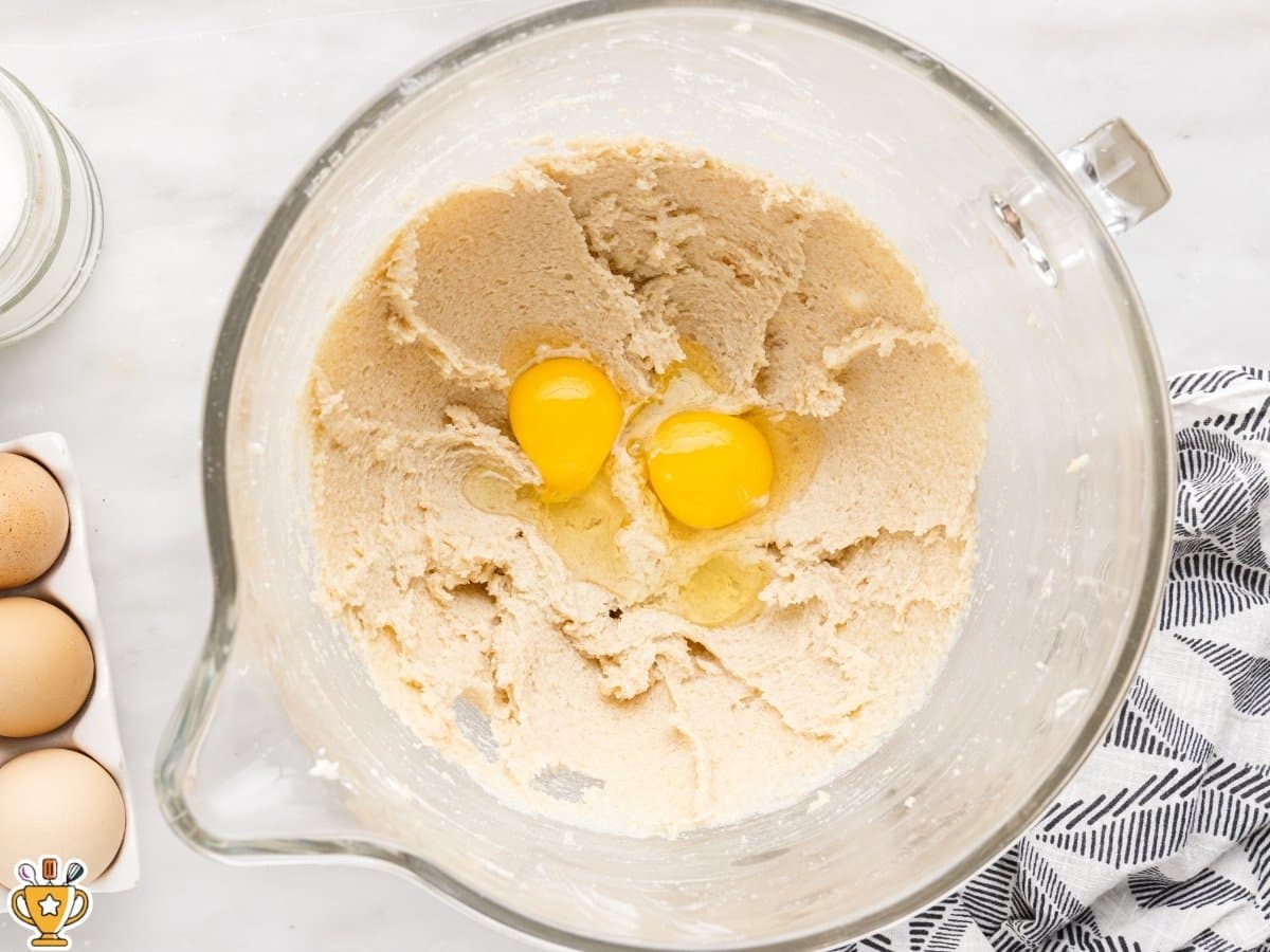 egg and egg yolk added to creamed butter and sugar mixture in a glass mixing bowl, overhead view on a white marble countertop