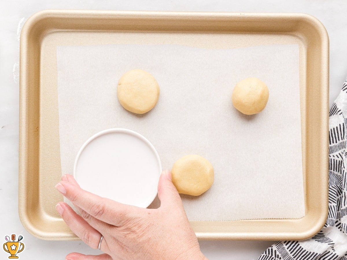 soft sugar cookie dough balls on a parchment-lined baking sheet being gently pressed flat before baking