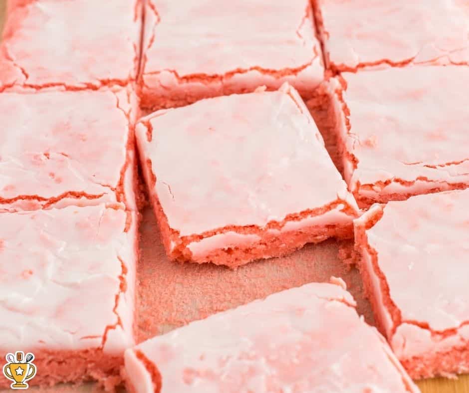 strawberry brownies cut into squares with pale pink glaze, one piece slightly lifted to show dense, soft interior on a parchment-lined pan