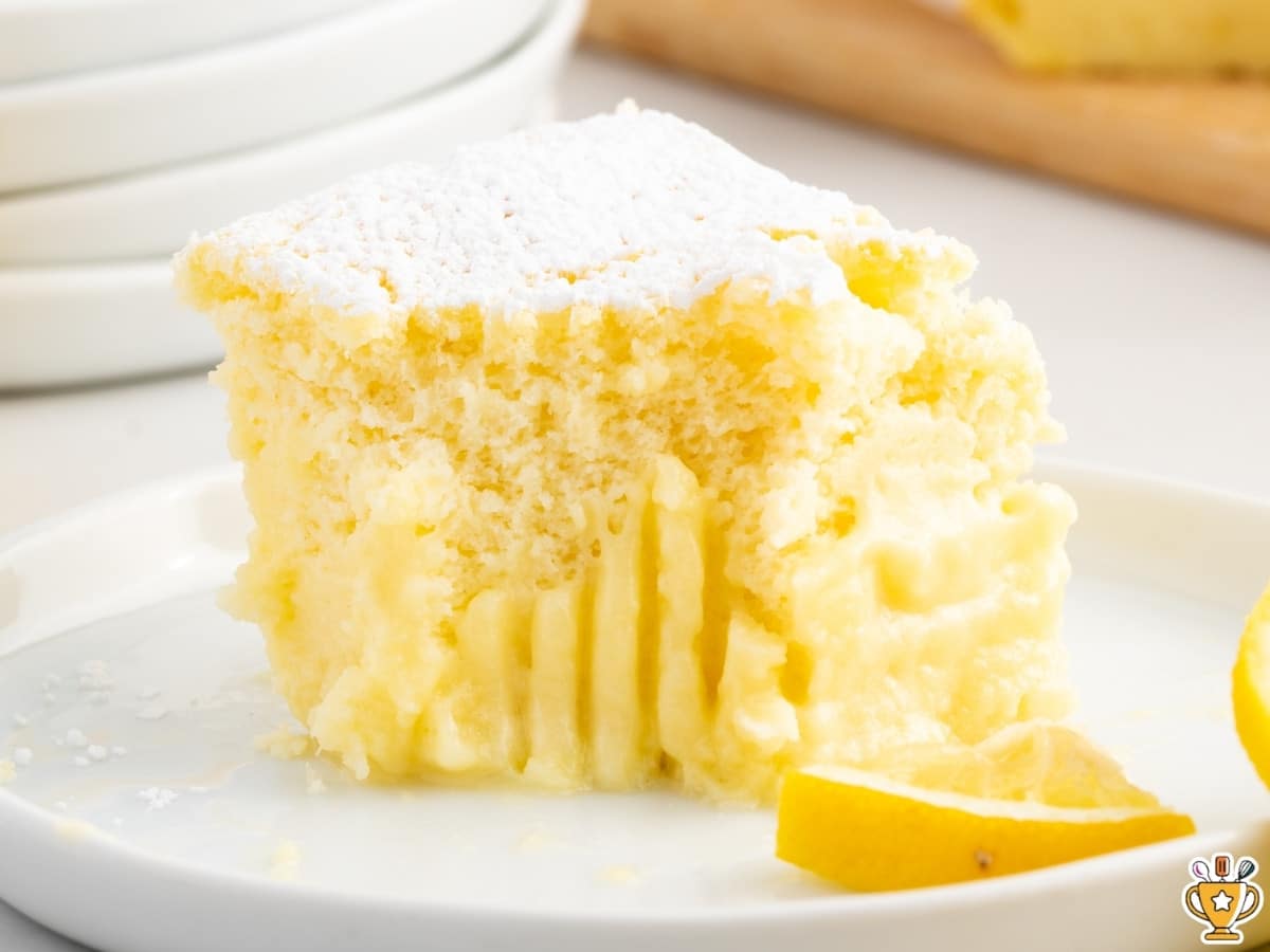 Close-up of a soft lemon custard cake slice with a light, airy sponge top and creamy custard layer underneath, dusted with powdered sugar and served on a white plate with a fresh lemon wedge.