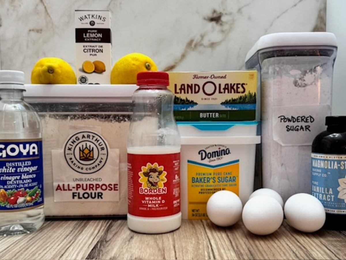Overhead view of measured ingredients for lemon custard cake, including flour, sugar, eggs, milk, butter, lemon juice, lemon zest, vanilla extract, lemon extract, vinegar, and powdered sugar arranged in bowls on a light surface.