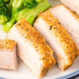 Close-up of sliced Parmesan crusted pork chops served on a white plate with steamed broccoli, showing the golden breadcrumb and Parmesan coating and the juicy center of each boneless pork chop.