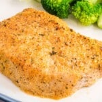 Single baked Parmesan crusted pork chop on a white plate with broccoli in the background, highlighting the crispy golden Parmesan breadcrumb coating on the boneless pork chop.