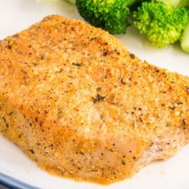 Single baked Parmesan crusted pork chop on a white plate with broccoli in the background, highlighting the crispy golden Parmesan breadcrumb coating on the boneless pork chop.