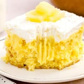 Slice of pineapple poke cake on a white plate topped with whipped topping and pineapple chunks, showing the moist yellow cake and creamy pineapple pudding filling.