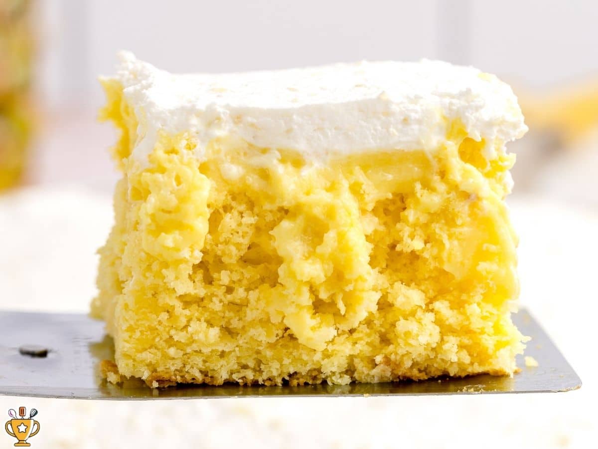 Close-up of a thick slice of pineapple poke cake on a metal spatula, showing the soft yellow cake, creamy pineapple pudding layer, and fluffy whipped topping.