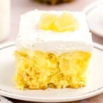 Square slice of pineapple poke cake on a white plate, topped with whipped topping and pineapple chunks, showing the soft yellow cake and rich pineapple pudding filling.