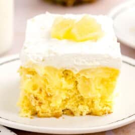 Square slice of pineapple poke cake on a white plate, topped with whipped topping and pineapple chunks, showing the soft yellow cake and rich pineapple pudding filling.