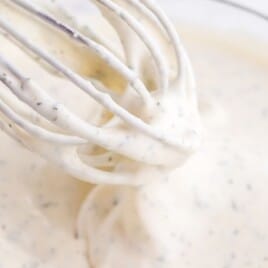 Close-up of creamy homemade ranch dressing being whisked in a clear glass mixing bowl, showing a smooth texture with herb flecks.