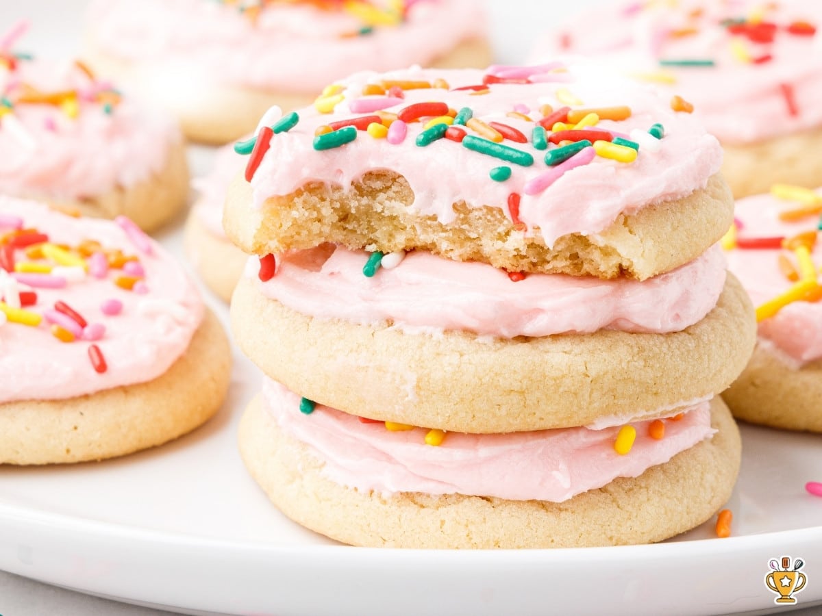 Soft sugar cookies stacked with pink frosting and rainbow sprinkles, one cookie broken open to show the soft tender center on a white plate