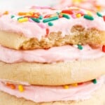 soft frosted sugar cookies stacked with pink frosting and rainbow sprinkles, one cookie broken open to show the soft tender crumb