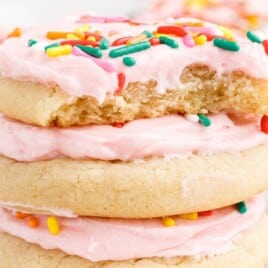 soft frosted sugar cookies stacked with pink frosting and rainbow sprinkles, one cookie broken open to show the soft tender crumb