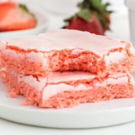 Close-up of frosted strawberry brownies cut into squares, showing the soft pink brownie layer under a crackly pale pink icing, with a bite missing out of the brownies.