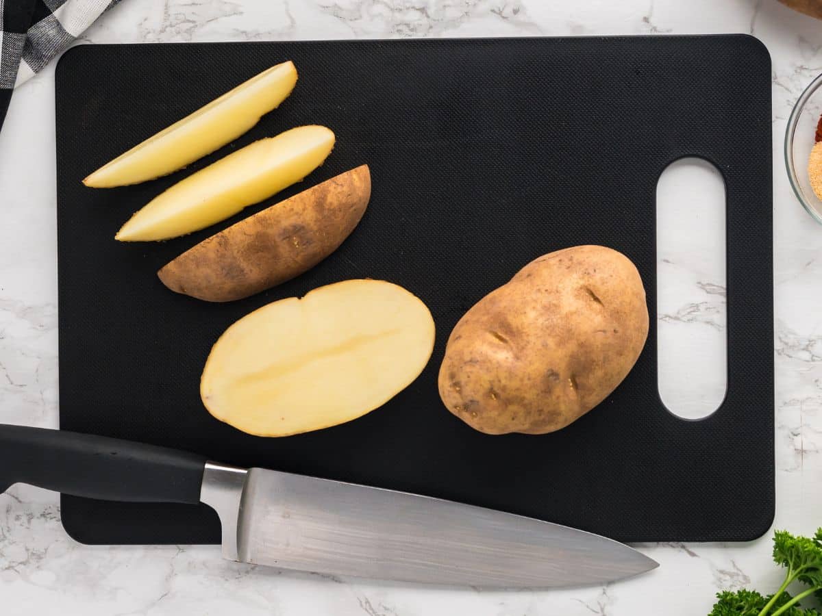 Russet potatoes on a cutting board being sliced into wedges with a chef&rsquo;s knife as the first step in preparing air fryer potato wedges.