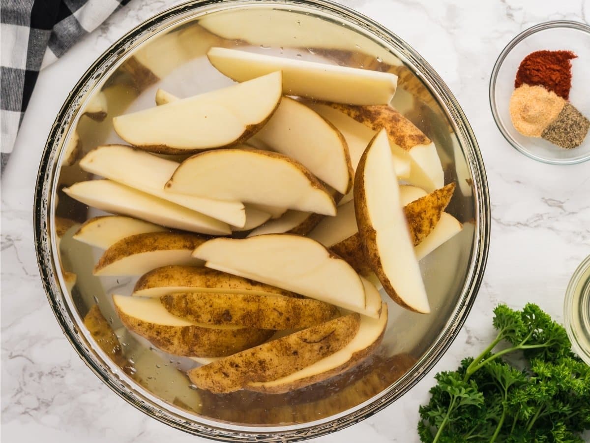 Freshly cut russet potato wedges soaking in a bowl of water to remove excess starch before air frying.