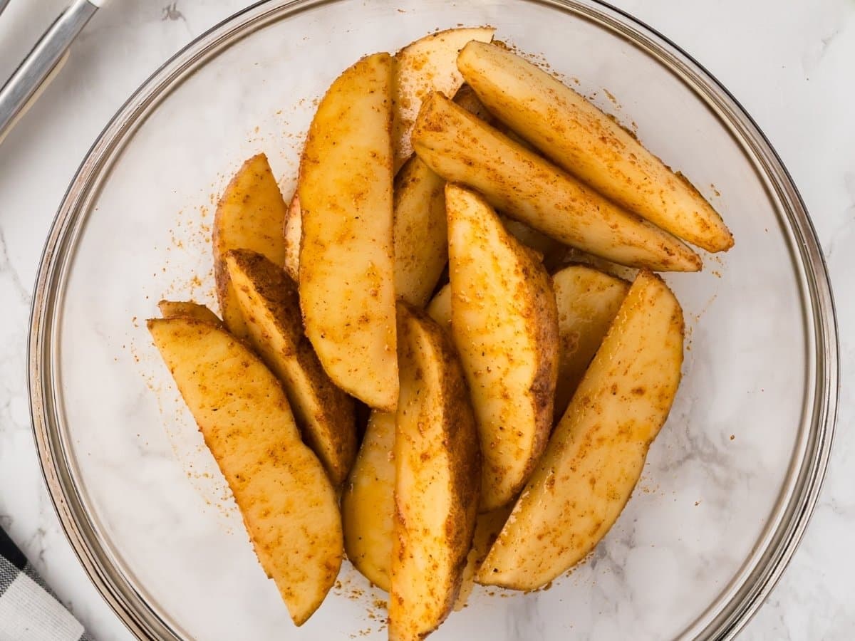 Potato wedges tossed with oil and seasoning in a bowl before cooking in the air fryer.