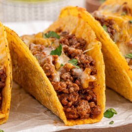 Close-up of baked hard shell tacos with crisp golden shells, filled with seasoned ground beef and topped with melted cheese, garnished with small pieces of fresh cilantro.