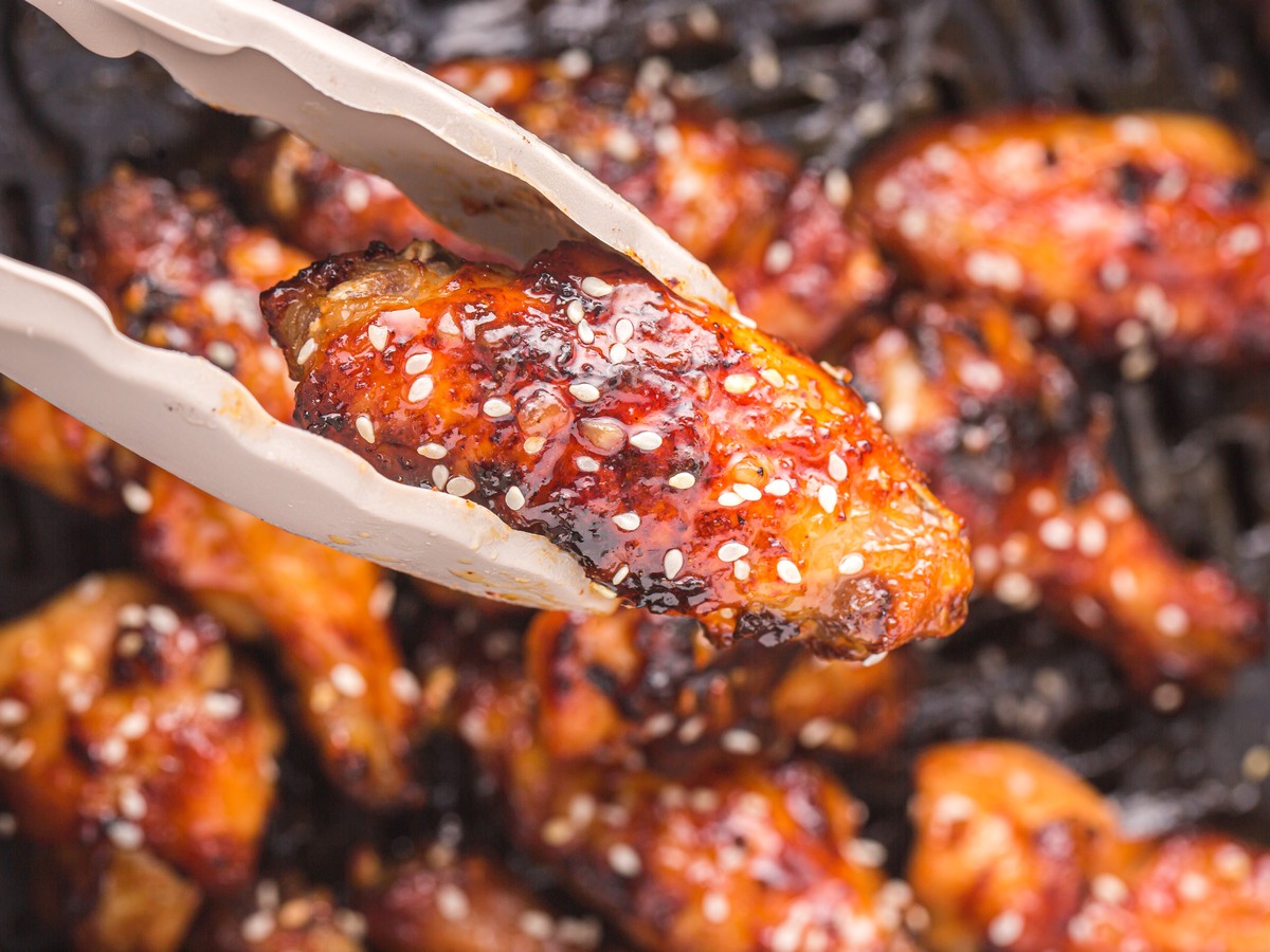 Close-up of tongs holding a glazed air fryer honey garlic chicken wing above the air fryer basket, showing sticky sauce, charred spots, and sesame seeds