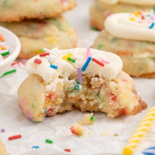 Featured image of Birthday Cake Cookies topped with swirls of white frosting and rainbow sprinkles on a white plate with milk and extra sprinkles.