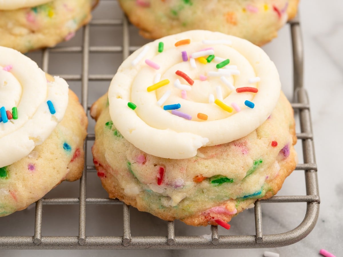 Overhead view of frosted Birthday Cake Cookies piled on a white plate with rainbow sprinkles, birthday candles, and extra sprinkles nearby.