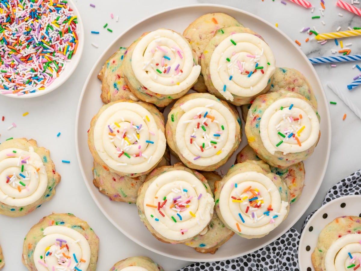Overhead view of frosted Birthday Cake Cookies arranged on a white plate, topped with swirls of vanilla frosting and rainbow sprinkles, surrounded by extra sprinkles and colorful birthday candles.