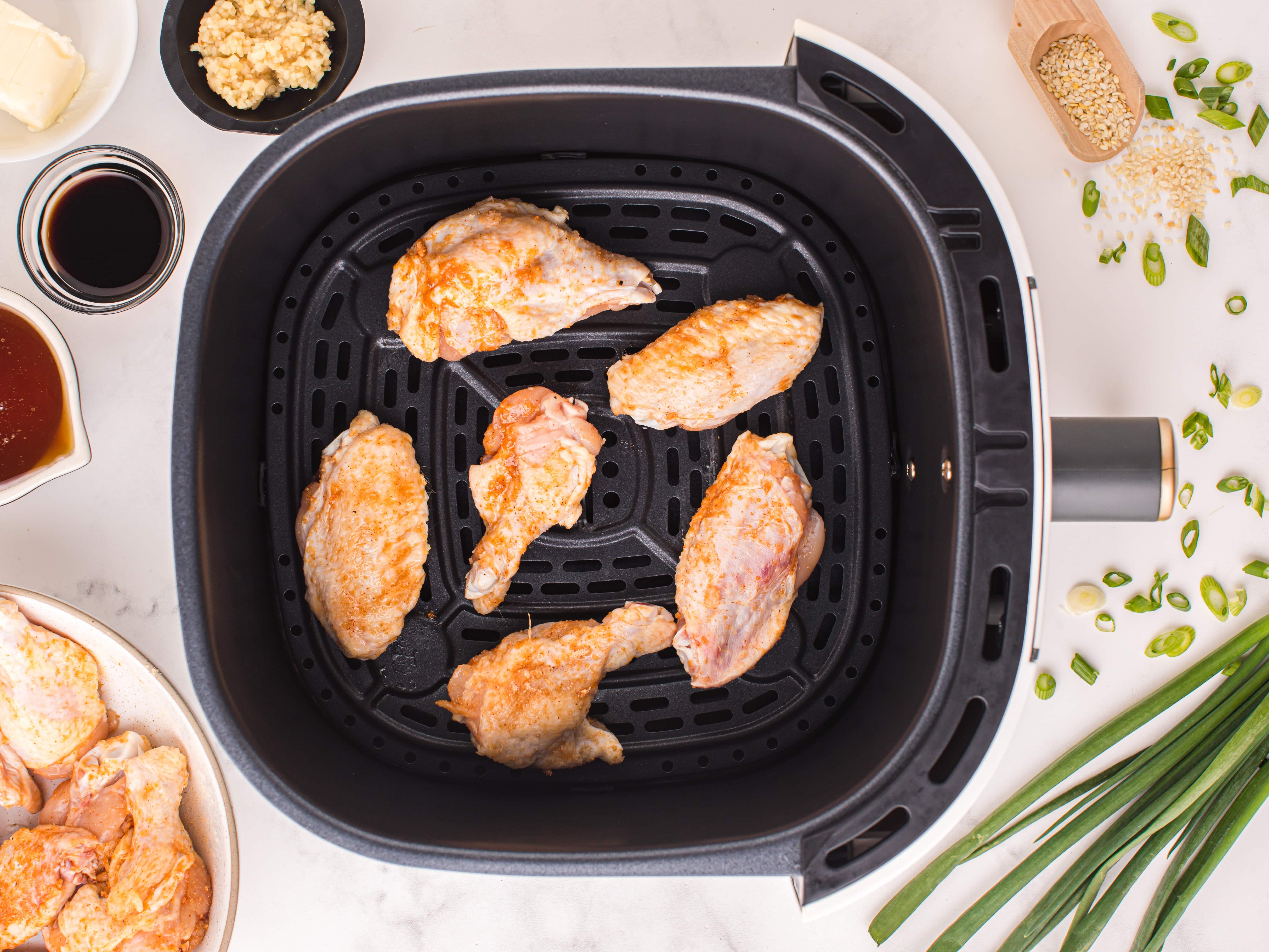 Chicken wings arranged in a single layer in an air fryer basket, surrounded by bowls of honey, soy sauce, garlic, butter, sesame seeds, and sliced green onions on a marble surface.