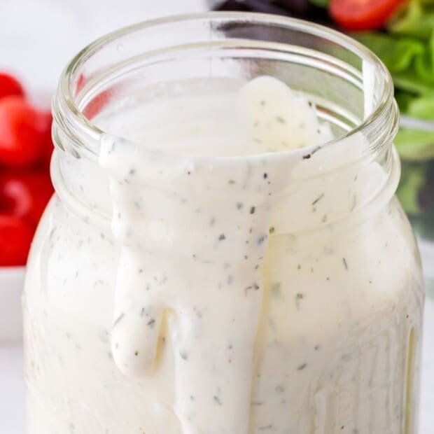 Creamy ranch dressing in a mason jar, thick and smooth with visible herb flecks, slowly dripping down the inside of the jar, with fresh vegetables blurred in the background.