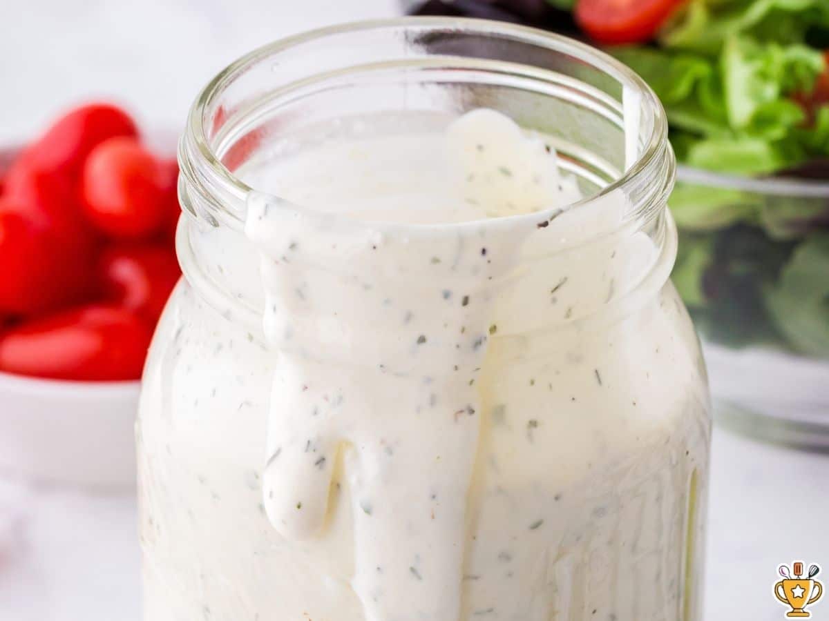 Creamy ranch dressing in a mason jar, thick and smooth with visible herb flecks, slowly dripping down the inside of the jar, with fresh vegetables blurred in the background.