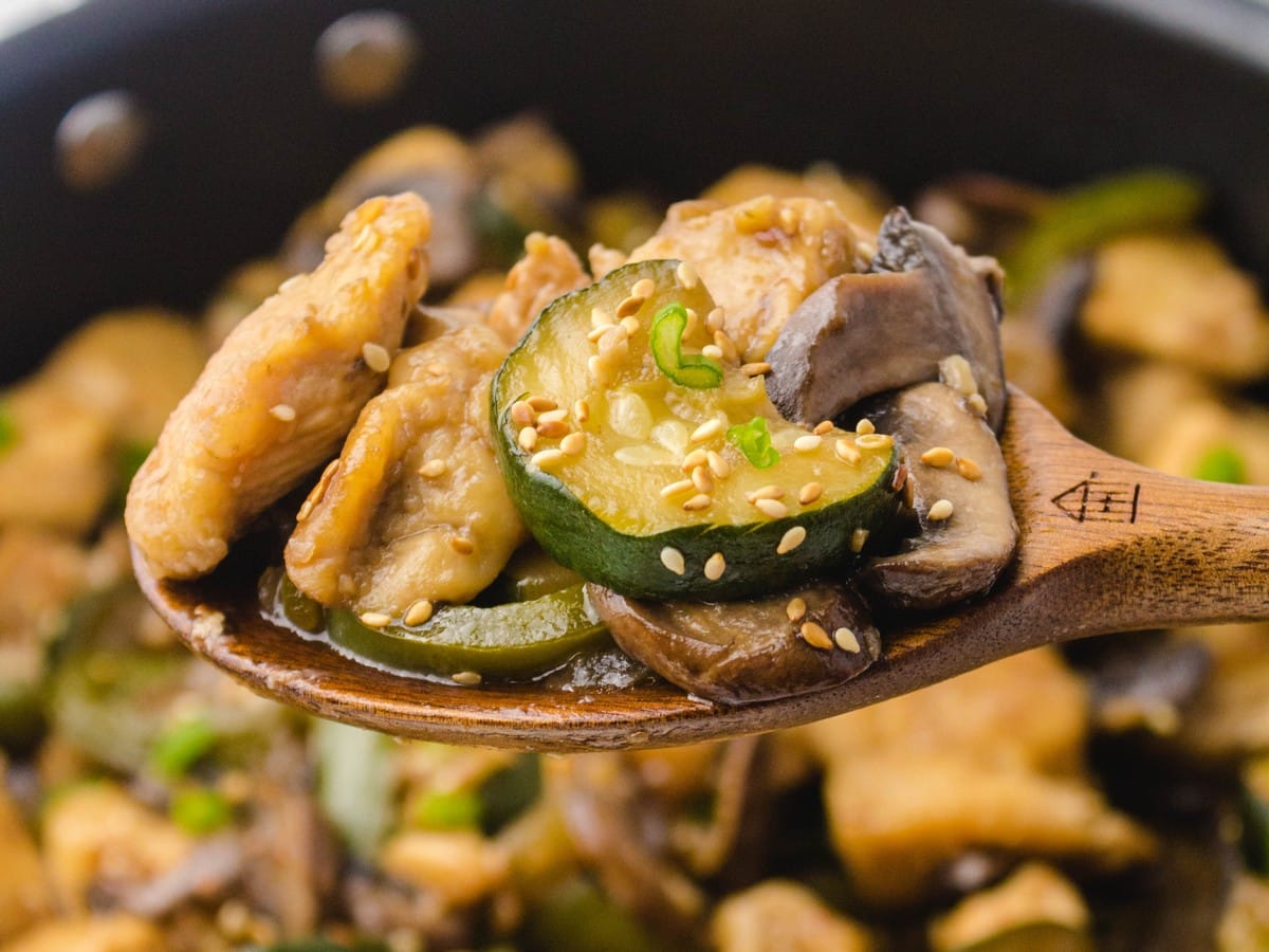 Wooden spoon lifting Panda Express mushroom chicken with juicy chicken, sliced mushrooms, zucchini, and sesame seeds over the skillet
