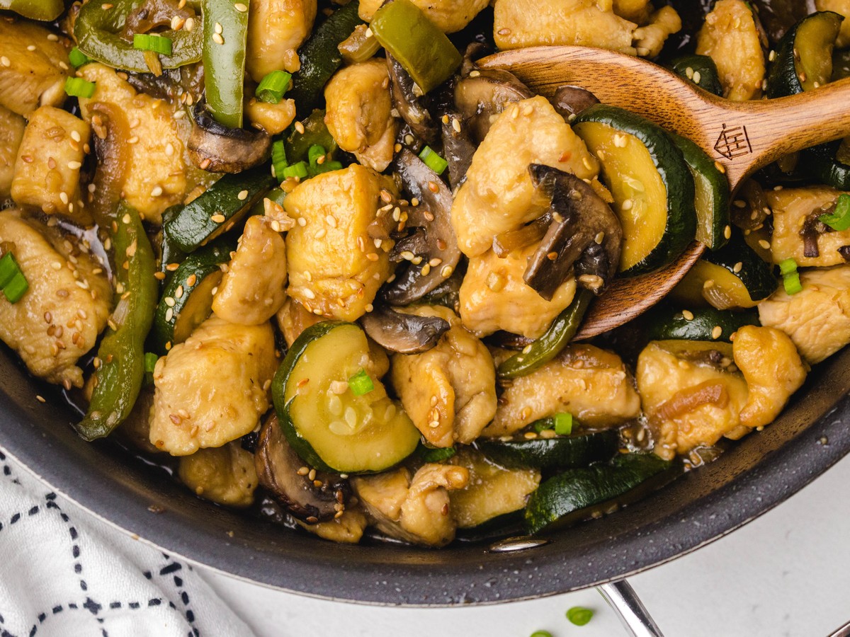 Overhead close-up of Panda Express mushroom chicken in a skillet with a wooden spoon, showing chicken breast pieces, mushrooms, zucchini, green peppers, and sesame seeds