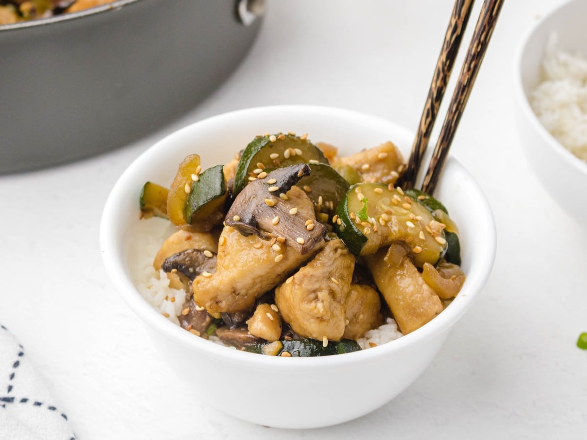 Bowl of Panda Express mushroom chicken served over white rice with chopsticks, topped with mushrooms, zucchini, chicken, and sesame seeds