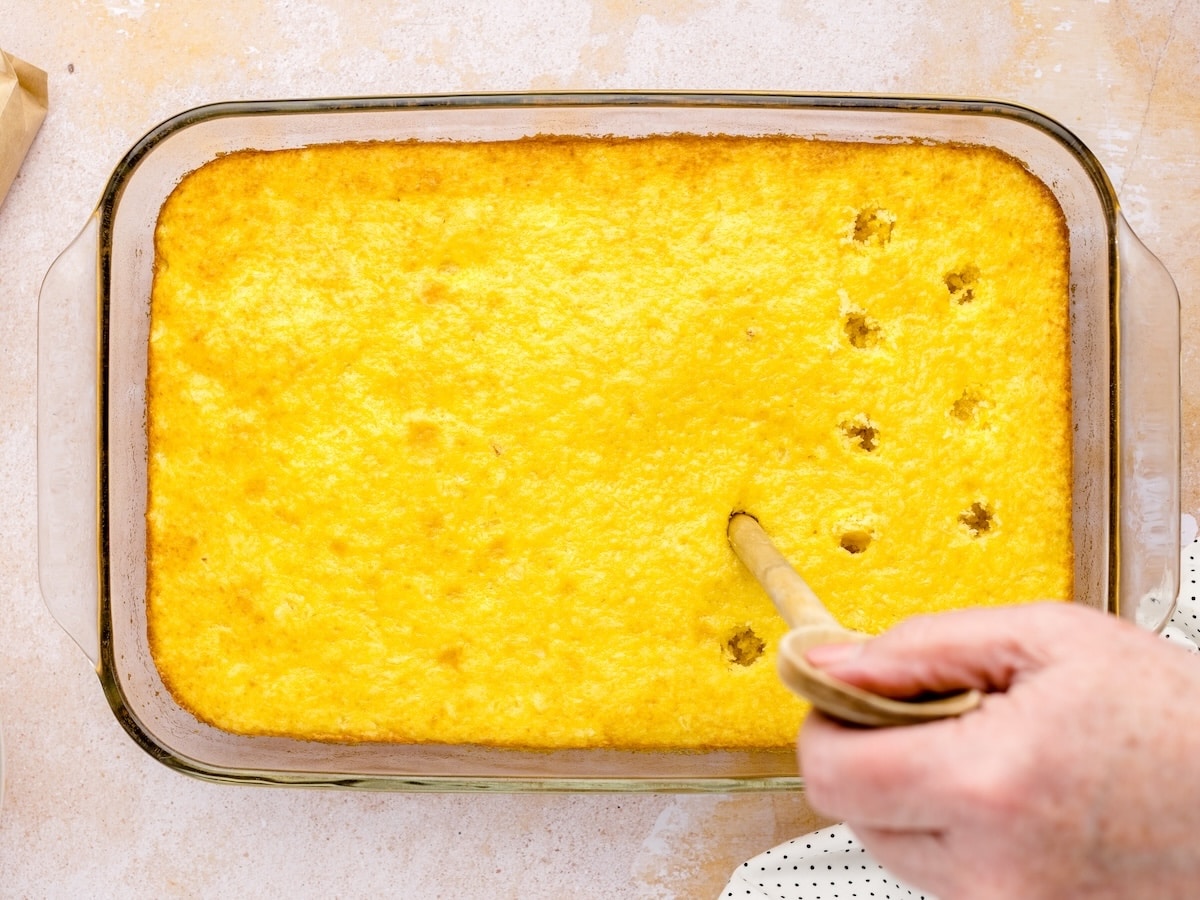 Poking holes in a baked pineapple cake using the handle of a wooden spoon, spaced about 1 inch apart to allow pudding to soak in evenly