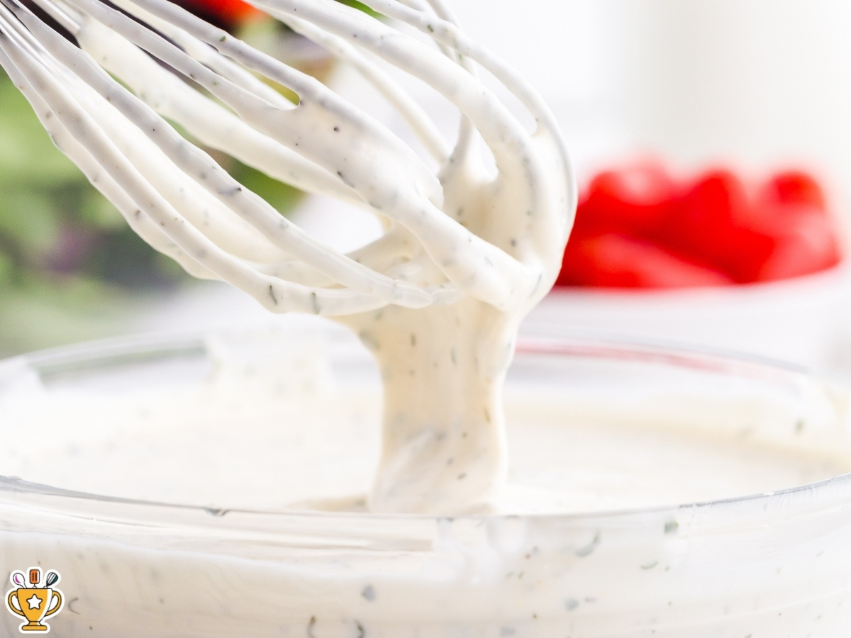Whisk lifting thick ranch dressing from a clear glass bowl, highlighting the creamy consistency and visible herb specks.