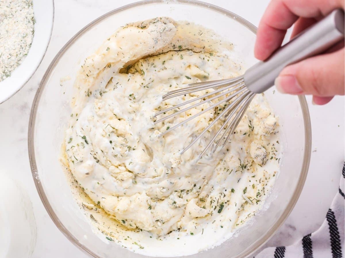 A hand is using a whisk to mix together ranch dressing ingredients in a medium glass bowl. 
