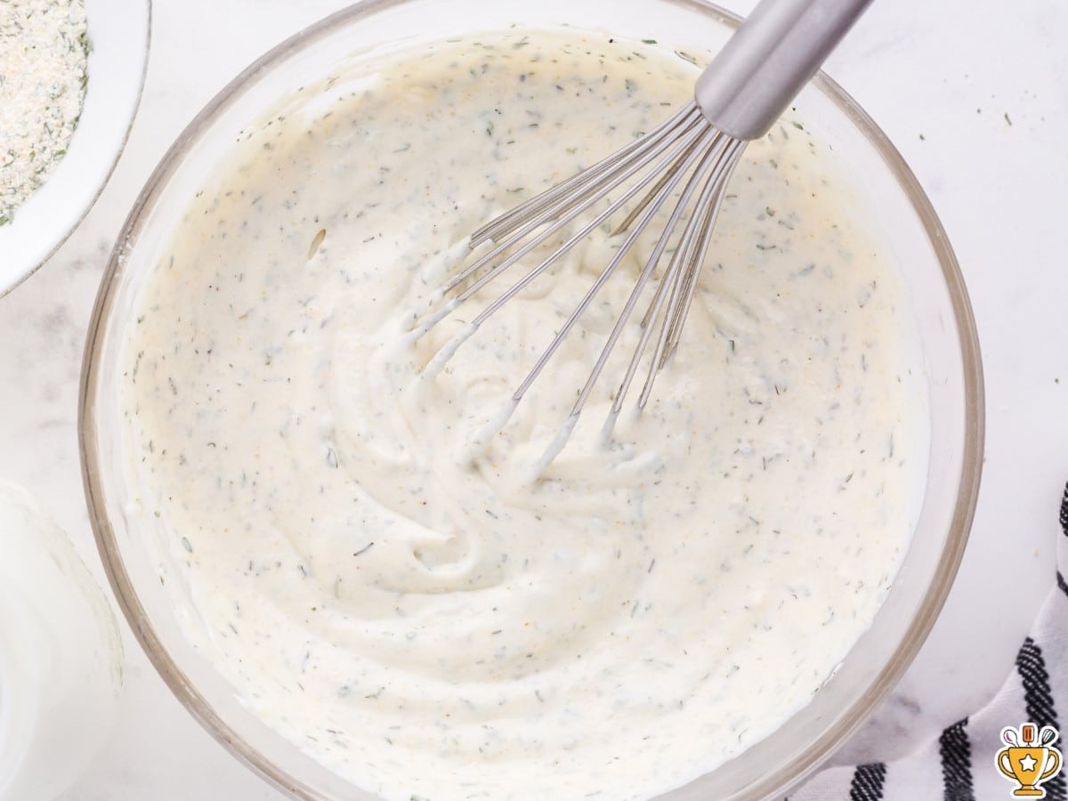 Smooth, fully mixed ranch dressing in a glass bowl, thick and creamy with evenly distributed herb flecks, being whisked to a uniform consistency.