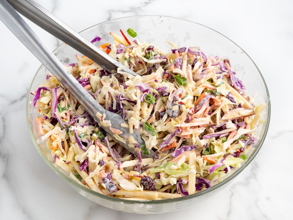 Apple slaw with cabbage, thinly sliced red apples, carrots, green onions, dried cranberries, and almonds being tossed in a creamy dressing with metal tongs in a glass bowl