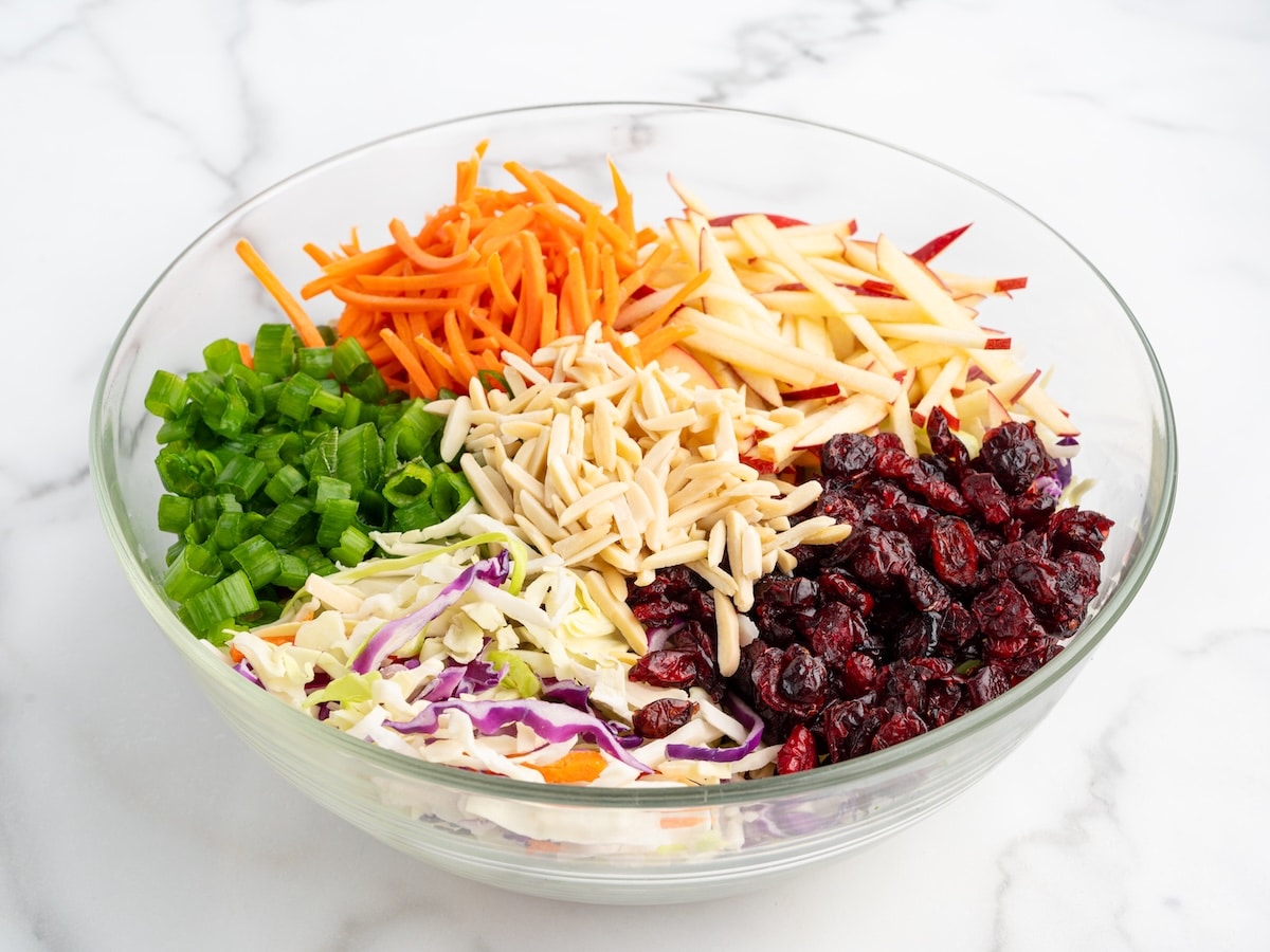 Coleslaw mix with sliced red apples, matchstick carrots, green onions, dried cranberries, and slivered almonds arranged in a glass mixing bowl before adding dressing.