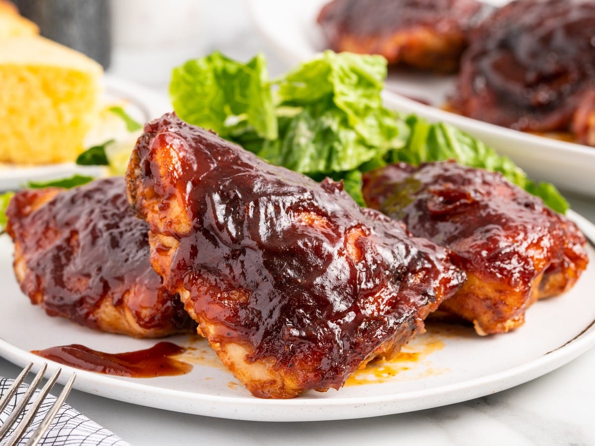 Baked Barbecue Chicken Thighs on a white plate coated in thick glossy BBQ sauce, served with green salad and cornbread, showing caramelized edges and juicy roasted chicken.
