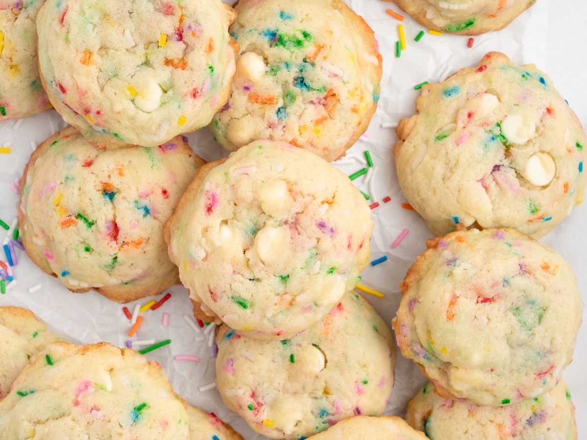 A overhead close-up of a pile of birthday cake cookies loaded with rainbow sprinkles and white chocolate chips on parchment paper.