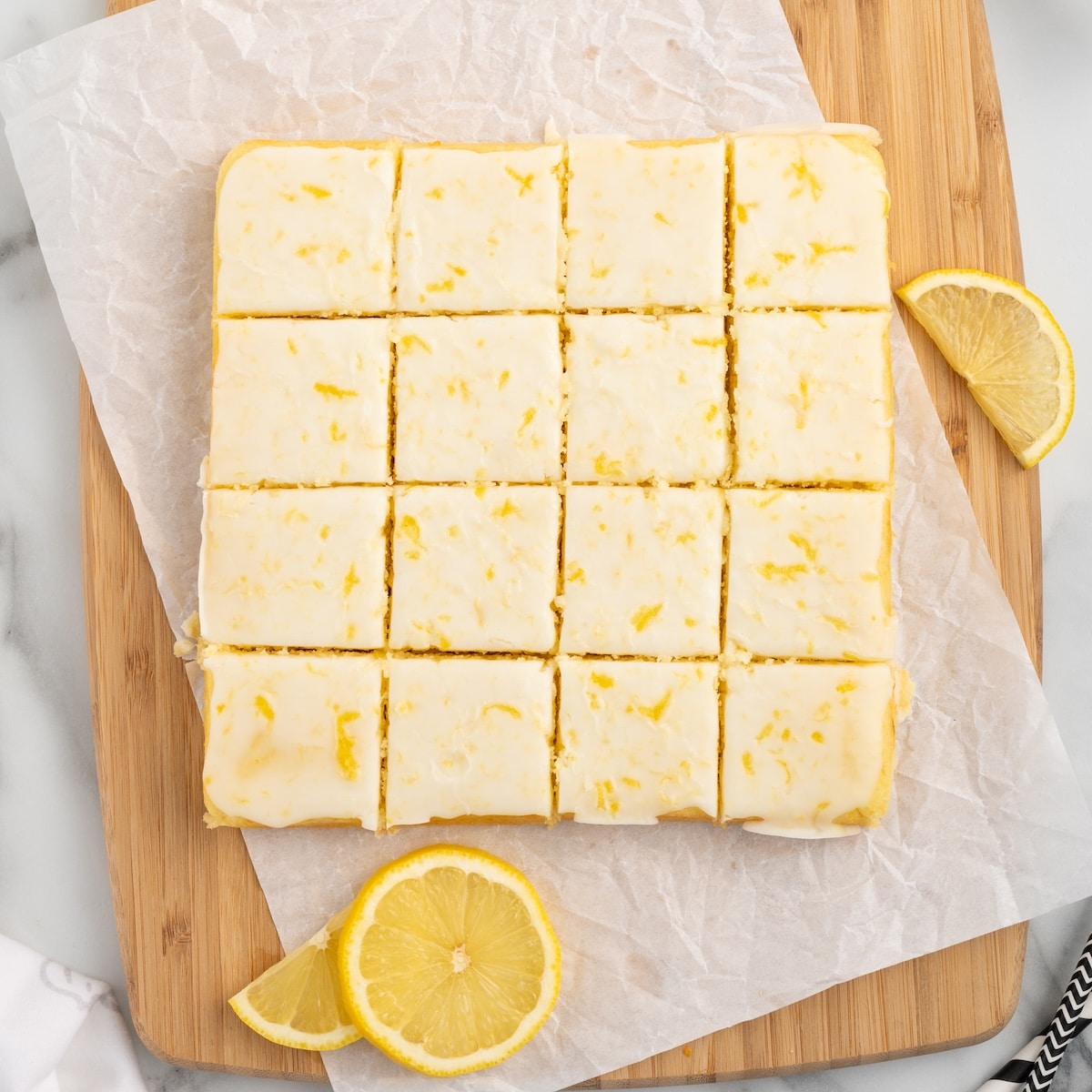 Sixteen glazed lemon blondies cut into squares on parchment paper on a wooden cutting board, with fresh lemon slices alongside.