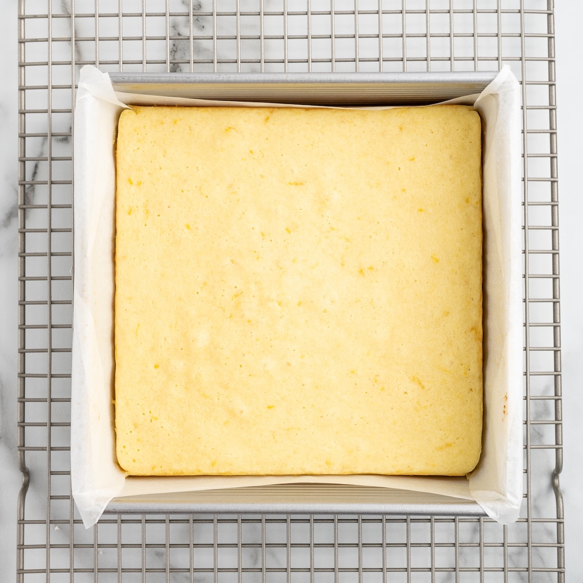Freshly baked lemon blondies in a parchment-lined pan on a cooling rack, showing the pale yellow color that tells you they are done.