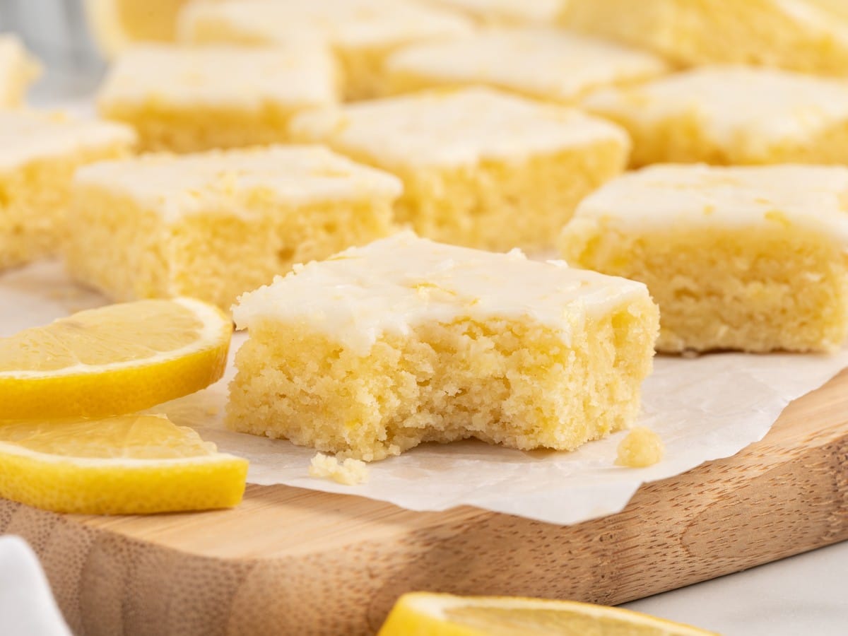 Tall stack of lemon blondies with soft, dense crumb and creamy lemon glaze, surrounded by lemon slices on parchment paper