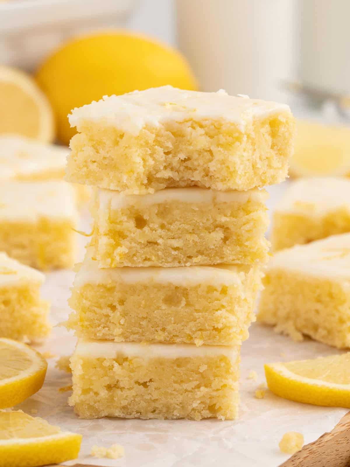 Close-up of lemon blondies stacked on a white plate, topped with smooth lemon glaze and flecks of lemon zest, with lemons in the background