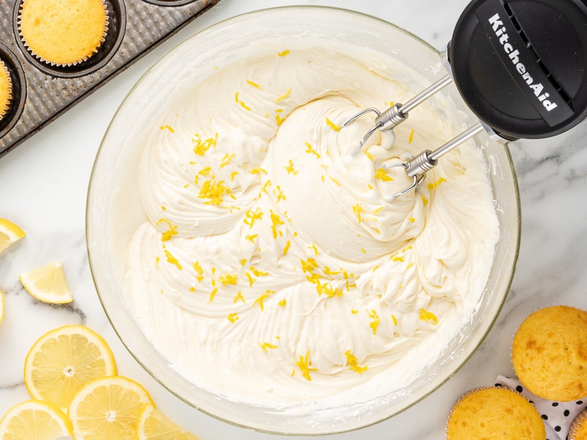 Overhead view of homemade Lemon Cream Cheese Frosting whipped until smooth and fluffy in a glass mixing bowl with lemon zest, electric mixer beaters, fresh lemon slices, and unfrosted cupcakes nearby.